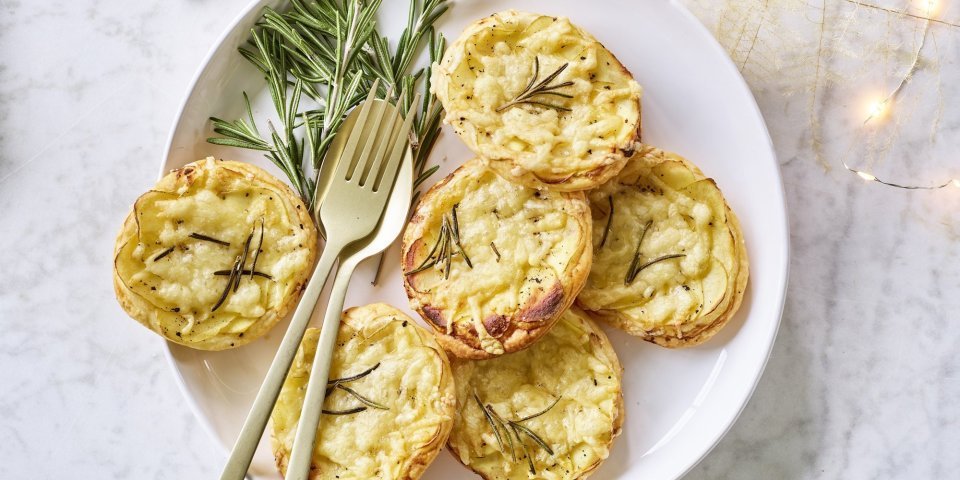 Taartjes met schijfjes aardappel en Belgische kaas liggen op een wit bord. Goud gestek ligt op het bord om meteen zo'n taartje op te scheppen. Er liggen enkele sterretjes en lichtjes langs om de feesttafel extra te laten blinken. 