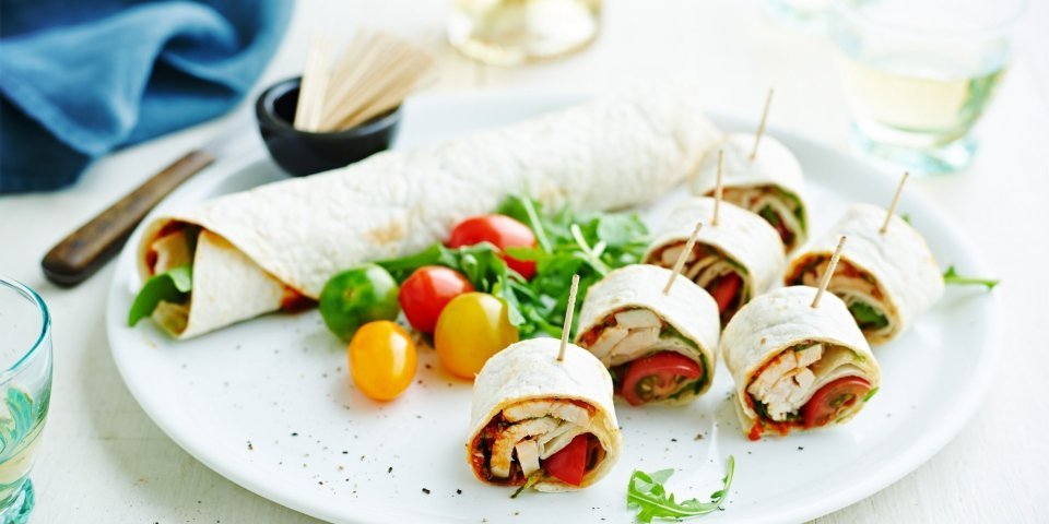 Op een bord ligt een rol wrap met groenten, wat kerstomaten en zes stukken wrap met een prikker in.