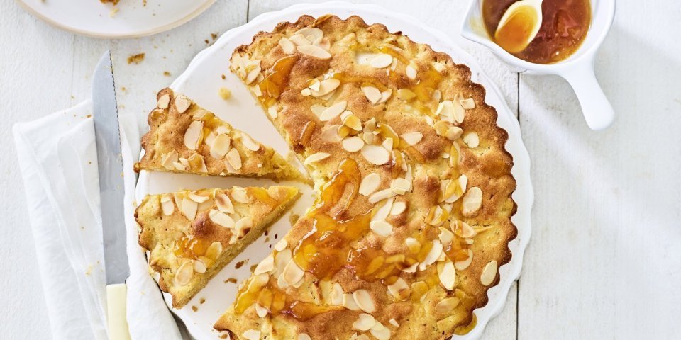 Appelcake met amandelen Goudgele appelcake staat op een houten witte tafel. De ronde cake is al voor een kwart aangesneden. Er is al een stukje uitgenomen en er staat een potje met abrikozengelei bij.