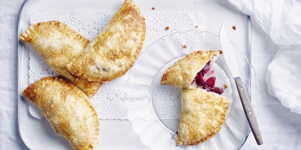 Er liggen 4 appelflappen klaargemaakt op een wit plateautje. Eén appelflap is opengesneden, door de combinatie te maken met blauwe bessen heeft de vulling mooi met appels een mooie rode kleur.