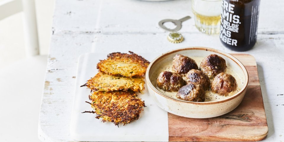 Balletjes in mosterdsaus met groenterösti Gebakken balletjes liggen in een diep bord op de mosterdsaus, de krokante groenterösti's liggen er langs. Er staat een flesje bier op tafel met een klein glaasje.