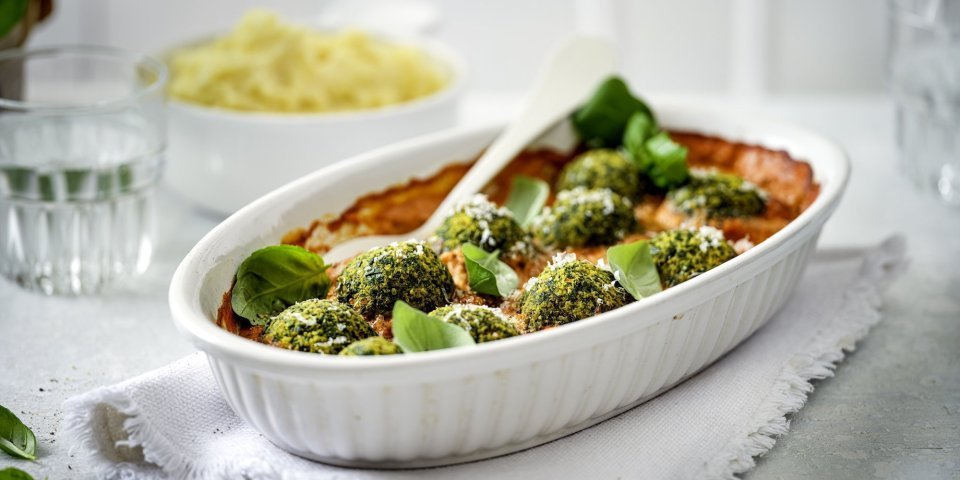 Een witte ovenschotel staat op tafel op een dikkere keukenhanddoek. De ovenschotel is gevuld met rode paprikasaus en hierop liggen de groene vegetarische balletjes van boerenkool. In de verte zie je ook nog een kommetje puree, klaar om aan tafel te gaan dus!