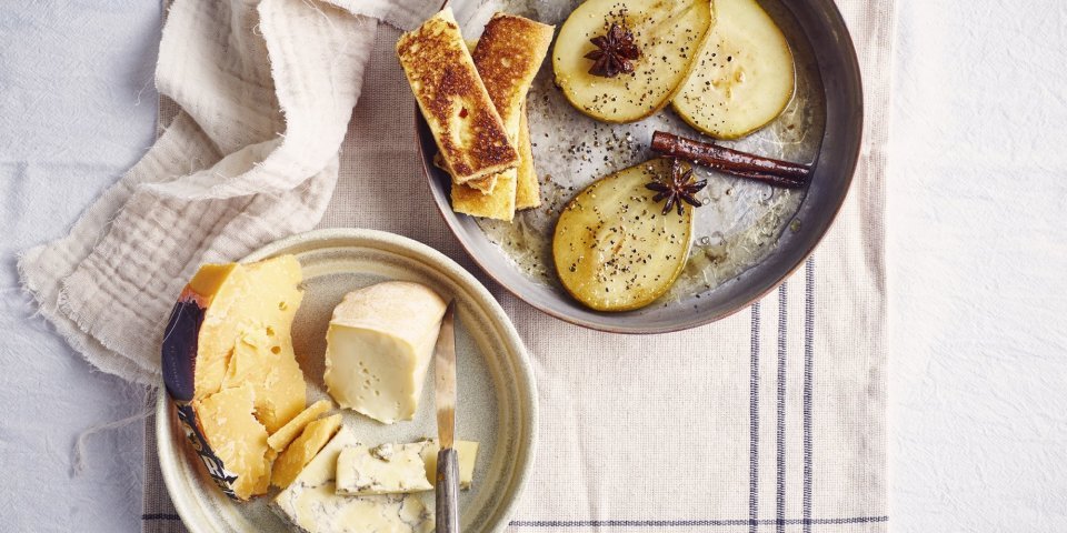 Briochetoast met gekarameliseerde peer, Belgische kazen en specerijensiroop Op tafel ligt een handdoek opengevouwen met hierop een bord en een pan. Op het bord ligt een verzameling Belgische kazen en een mes, in de pan liggen gekaramiliseerde peren, safraanstokje, steranijs en briochetoastjes.