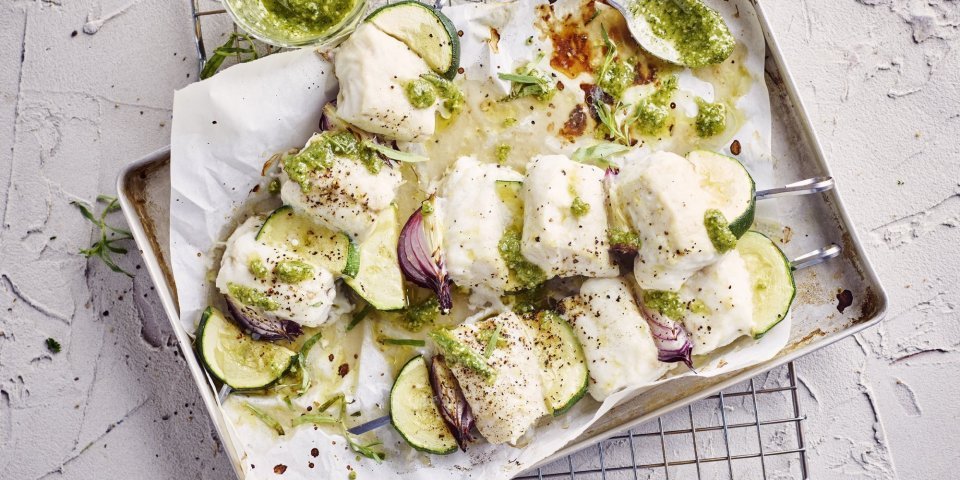 Brochette van heek met courgette, rode ui en dragonpesto Klaargemaakte brochettes liggen in een grijze ovenschaal te blinken. De spiesjes bestaan uit stukjes heek, courgette en rode ui. Er staat een glazen potje naast met huisgemaakte dragonpesto.