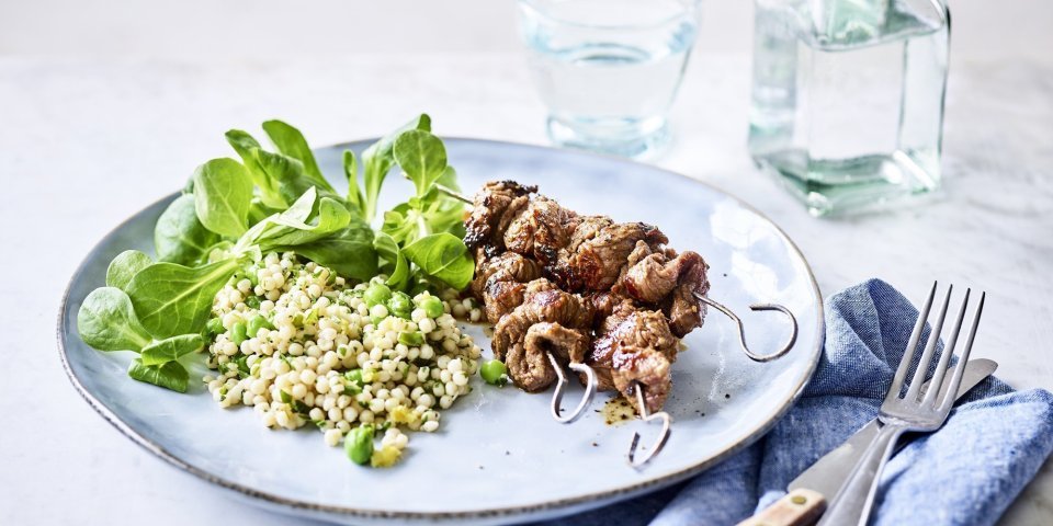 Brochette van rundvlees met salade van parelcouscous, erwtjes, verse kruiden en veldsla Op een lichtblauw groot bord liggen 3 gegrilde brochettes, we serveren ze met een couscoussalade van erwtjes en verse kruiden. Naast het bord ligt een blauwe servette met bestek langs. Er staat ook een glaasje water bovenaan het bord met een flesje water naast.