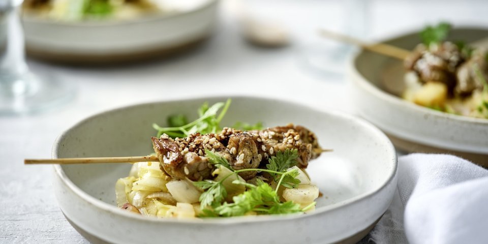 Er staan verschillende aperetiefbordjes op tafel met een schepje gestoofd witloof en appel. Hierop ligt een klein brochette van rundvlees en afgewerkt met kervel. 