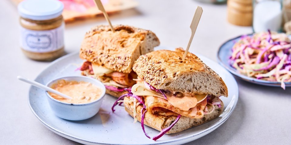 Broodje Breydelspek met koolsla, kaas en pittig sausje Er liggen twee broodjes op een licht blauwgrijs bordje. De broodjes zijn belegd met breydelspek, koolsla van rode kool en spitskool, een sneetje kaas en homemade saus.