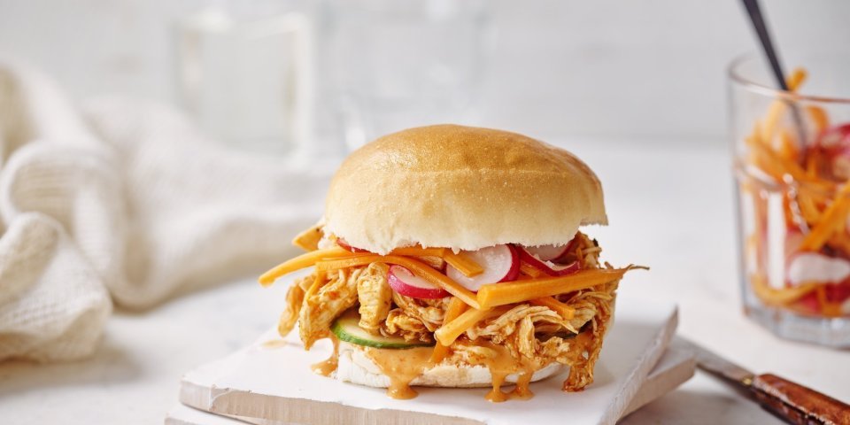Broodje pulled chicken met chilimayonaise Een heerlijke pistolet ligt op een houten snijplankje. Het is opengesneden en gevuld met de boterzachte kip, zoetzure groentjes en chilimayonaise. In de achtergrond ligt nog een servette om je smosvlekken snel op te drogen.