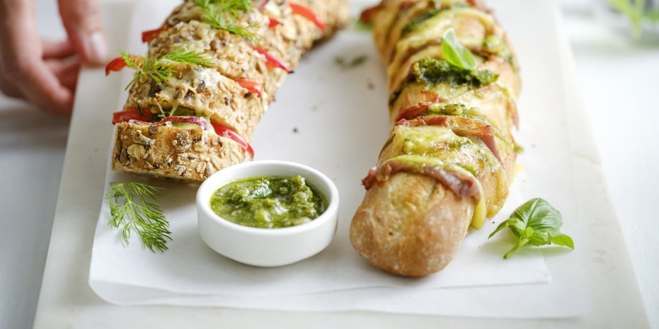 Broodjes op de BBQ Twee broodjes liggen op een snijplank. Je ziet dat het twee versies zijn, nl een broodje met groene pesto, sneetjes ham en kaas. En dan nog een versie met roomkaas, paprika en rode ui. Extra pesto staat in een potje bij de broodjes, voor wie wat extra wil. Een vrouw zet de plateau of snijplank op tafel, haar handen zijn nog in beeld.