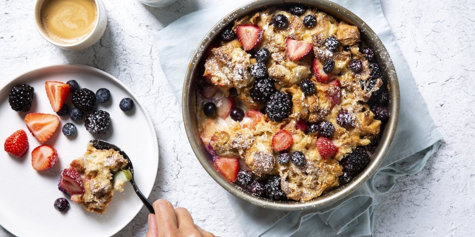 Broodpudding met rabarber, aardbeien, bramen en blauwe bessen Homemade broodpudding met rabarber en zomerbessen staat op tafel. Een vrouwelijke gast schept haar een portie uit om volop te genieten van dit dessert met een kopje koffie.