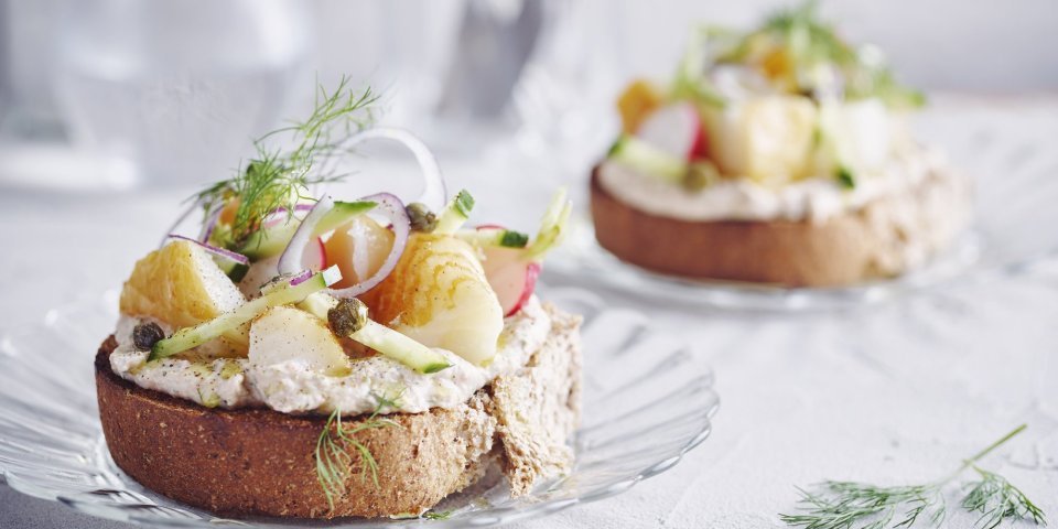 Bruschetta met bonenpuree, gerookte heilbot, radijzen en komkommer Een goedgevulde bruschetta ligt op een klein glazen bordje en staat op tafel. De bruschetta werd afgewerkt met een takje dille. Achteraan de tafel staat een kan met water en bestek om deze toast te verorberen.