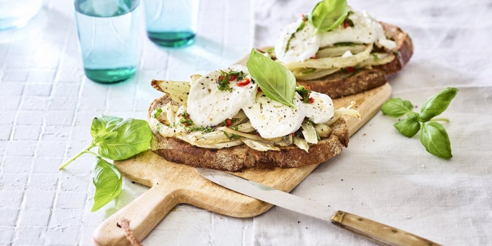 Bruschetta met geroosterde venkel en mozzarella Een rijkbelegde toast met venkel en mozzarella liggen op een houten plank. Boven deze plank staan twee blauwe glazen gevuld met water. Het gerechtje is afgewerkt met enkele basilicumblaadjes.