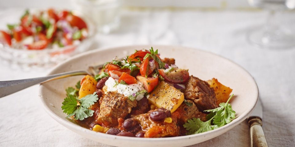 Chili con carne van stoofvlees met tomatensalsa en zure room Stoofvlees gemaakt met de smaken van een chili con carne, heerlijk! Er is een portie uitgeschept in een diep bord en staat op een gedekte tafel. In de achtergrond staat de tomatensalsa en zure room om het gerechtje helemaal af te werken.