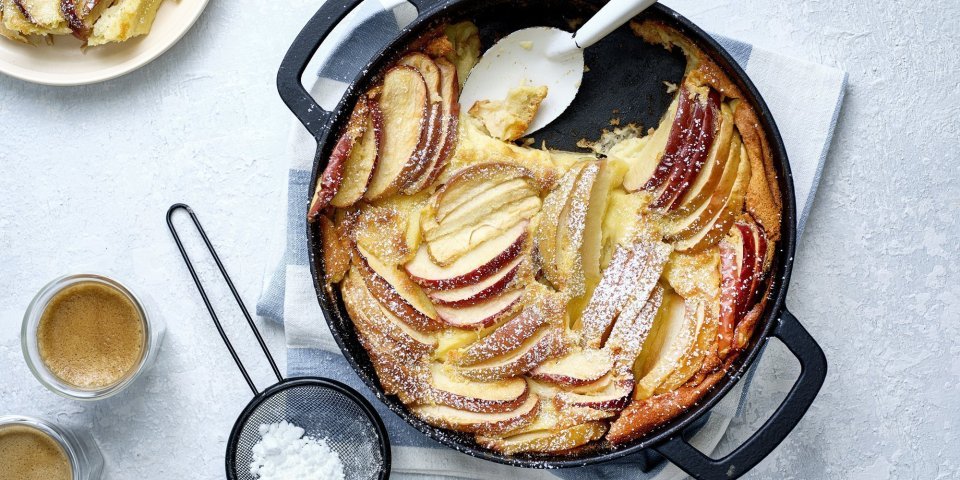 Clafoutis van appel Een clafoutis van appelstukkjes staat op het aanrecht in een zwarte fonte schaal. Er is al een portie uitgeschept en staat op een bordje naar de pot. Er is ook bloemsuiker over het dessert gestrooid en de koffie is al uitgeschonken.