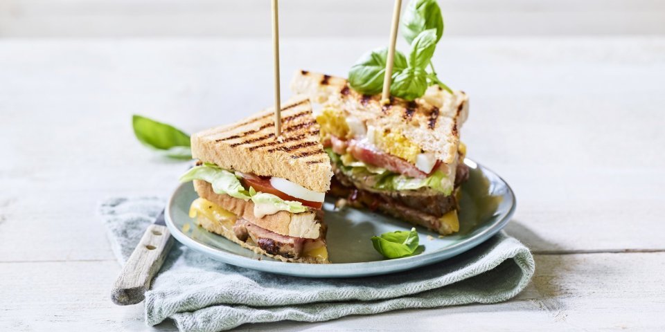 Goedgevulde club sandwich met lamsvlees en rauwkost. Gepresenteerd op een bord met middenin een prikker, klaar om op te eten. 