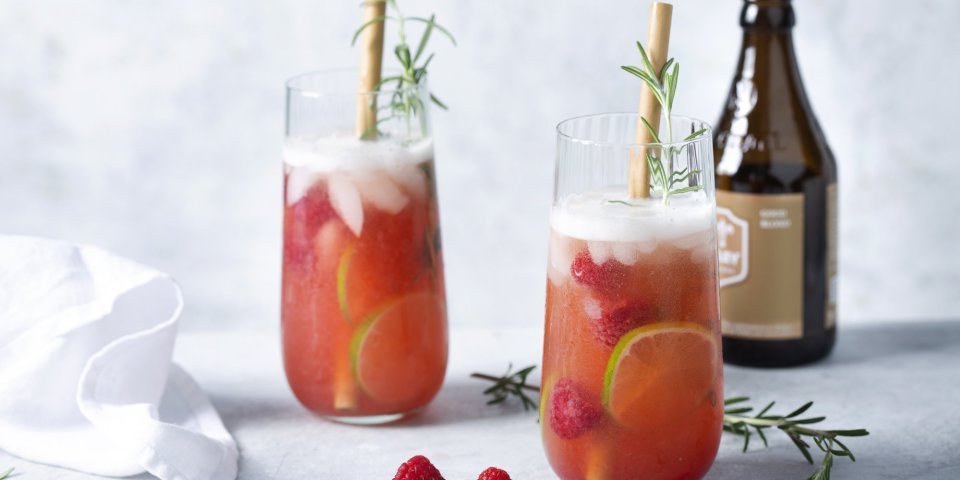 2 cocktails staan uitgeschonken in grote glazen. Deze cocktail op basis van bier en gin werd afgewerkt met frambozen en rozemarijn.