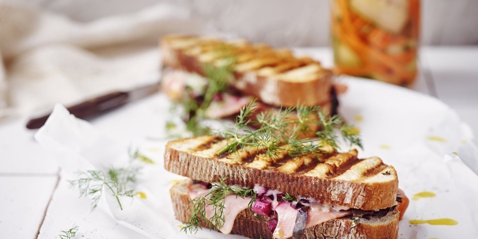 Twee croques liggen op het aanrecht in de keuken op een wit boterpapiertje. In de achtergrond staan de gepekelde groentjes (wortel en komkommer) in een bokaal. De tosti's zijn mooi gegrild in streepjes.