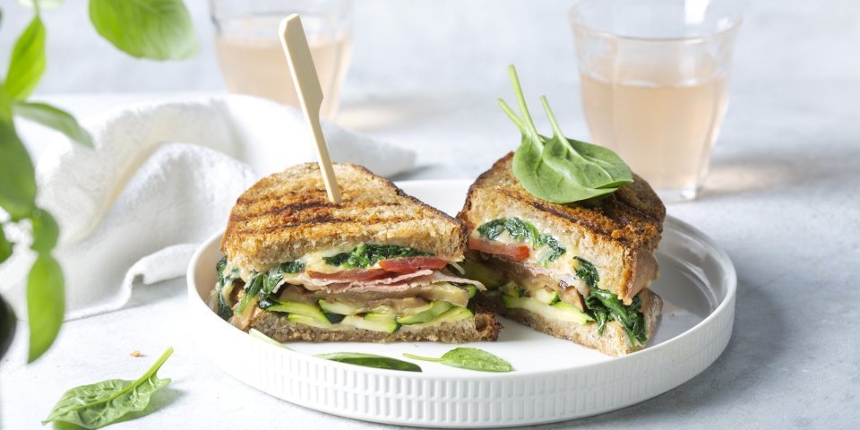 Croque met kaas, spinazie en gegrilde courgette en aubergine Een aangesneden croque ligt op een wit bord. De croque zit boordevol groentjes, en zit vast met een houten prikker. Achteraan de foto staan 2 glaasje licht roze huisgemaakte limonade.