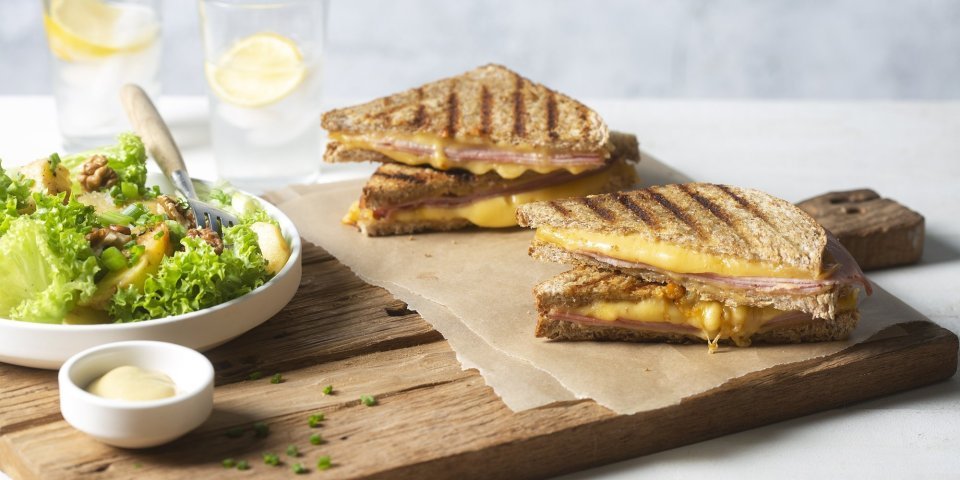 Een krokant gebakken croque monsieur ligt aangesneden op een houten snijplank. Lang staat een salade in een diep wit bord met een potje mayonaise voor te dippen. 