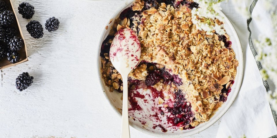 Crumble met havermout en braambessen in een witte ronde ovenschaal. Er is al een portie crumble verdwenen uit de ovenschaal en er liggen enkele braambessen langs. Om nog wat extra sfeer toe te voegen zijn er witte fijne bloemetjes rechts in beeld.