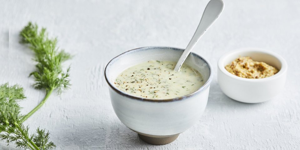 Dillesaus met mosterd De dillesaus is geserveerd in klein porselein potje, er zit een witte lepel in en een takje dille langs.
