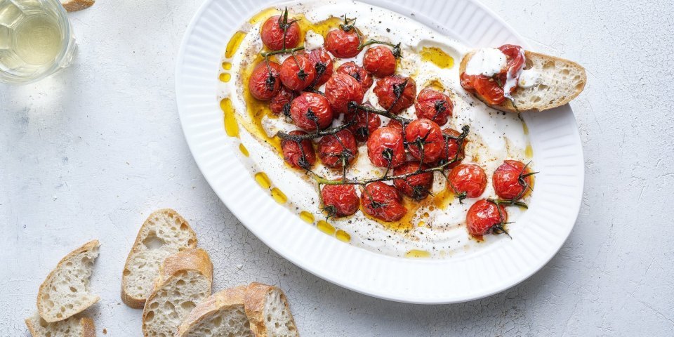 Dip van yoghurt met geroosterde tomaten Een groot ovaal bord is gevuld met de romige yoghurt waarop de dieprode kerstomaatjes bovenop liggen. Er liggen enkele sneetjes stokbrood naast om te dippen. Er staan ook twee glaasjes witte wijn in beeld.