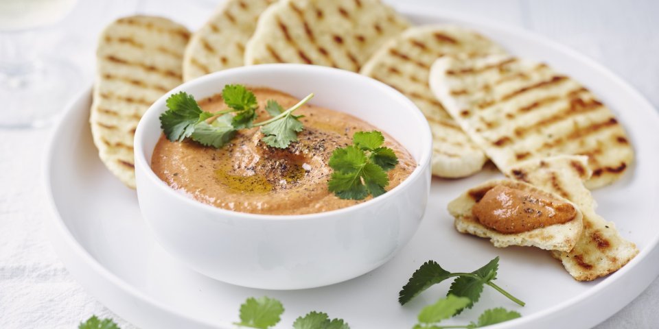 Homemade paprikadip zit in een wit schaaltjes. Het naanbrood ligt er langs op een wit bord. Het gercht is afgewerkt met korianderblaadjes.