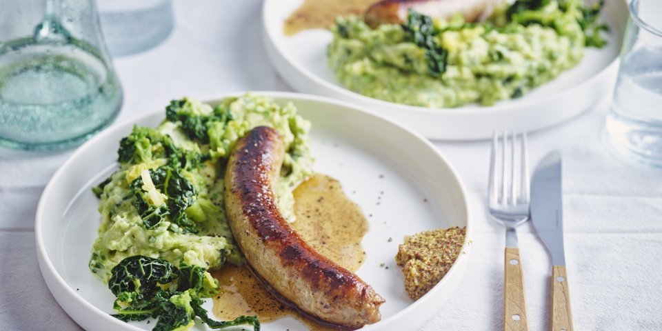 Erwtenpuree met savooi, braadworst en graantjesmosterd Op tafel staan twee borden gevuld met een schep erwtenpuree en savooikool. We serveren hem als een echte Vlaamse klassieker met een braadworst en een schep mosterd.