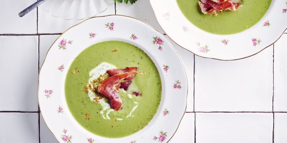 Erwtensoep met krokante ham 2 porties erwtensoep staan klaar op het werkblad van de keuken. De erwtensoep heeft een mooie groene kleur die mooi contrast geeft in de witte borden. De soep is afgewerkt met peterselieyoghurt, krokante ham en gefruite uitjes.