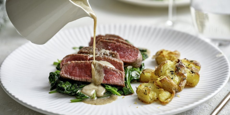 Een wit bord staat op tafel met hierop de heerlijke krokante aardappel en de saignant gebakken filet pur op een bedje van spinazie. Er komt een sauskannetje in beeld die saus over het vlees giet. De tafel is gedekt voor twee met de perfecte maaltijd.