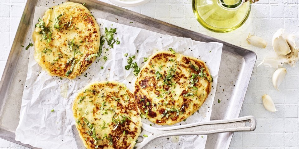 Flatbread van aardappel Drie broodjes zijn gebakken, en wat voor een! Flatbread gemaakt van een restje aardappelpuree liggen te blinken op een grijze bakplaat met wit boterpapiertje op. Verse kruiden liggen klaar om af te werken.