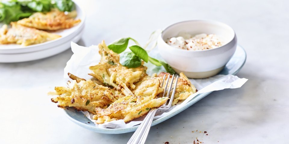 Fritters van aardappelen en Belgische kaas Meerdere fritters liggen op een licht blauw bordje vergezeld van een blaadje veldsla en het dipsausje op basis van yoghurt en specerijen.