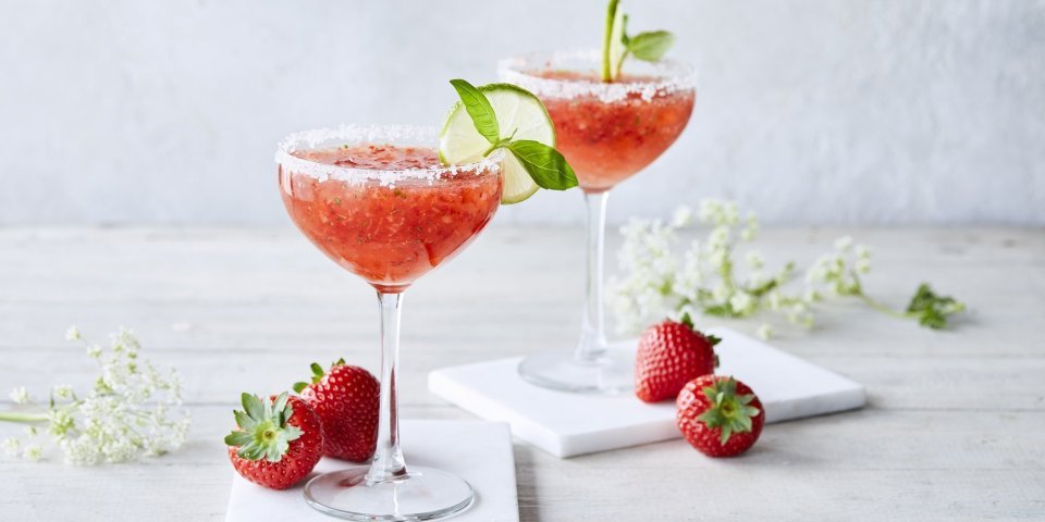 Cocktail time! Wij serveren een frozen daiquiri met verse aardbeien en basilicum. We presenteren hem in een wijnglaasje met suikerrandje, schijfje limoen en basilicumblaadje. Er liggen enkele aardbeien langs samen met witte bloemetjes. Schol! 