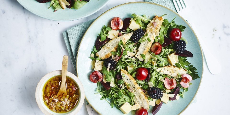 Fruitige salade met rode biet en gegrilde kip Een gemengde salade met sla, kersen, bramen en gekookte kipfilet gedresseerd op een lichtblauw bord. De dressing is er naast gezet in een keramieke potje met houten lepeltje in, de dressing bestaat uit sjalot, rode wijazijn, olijfolie en peper en zout.