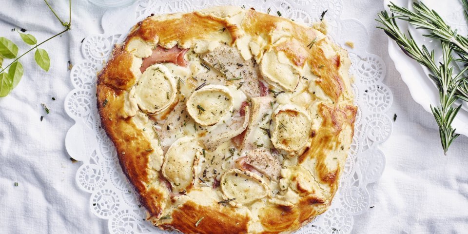 Galette met geitenkaas, peer en ham Een plaattaart of galette ligt op een rond taartpapiertje, klaar om aangesneden te worden. Er liggen enkele takjes rozemarijn langs voor de afwerking.