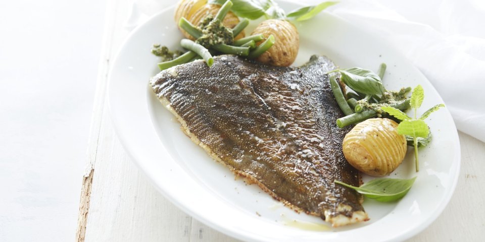 De gebakken pladijs op de graat ligt op een groot wit ovaal bord. Wij serveren hem met boontje langs en enkele hasselback potatoes. Het bord staat op een witte houten tafel en de salsa verde staat apart in een potje boven het bord.  