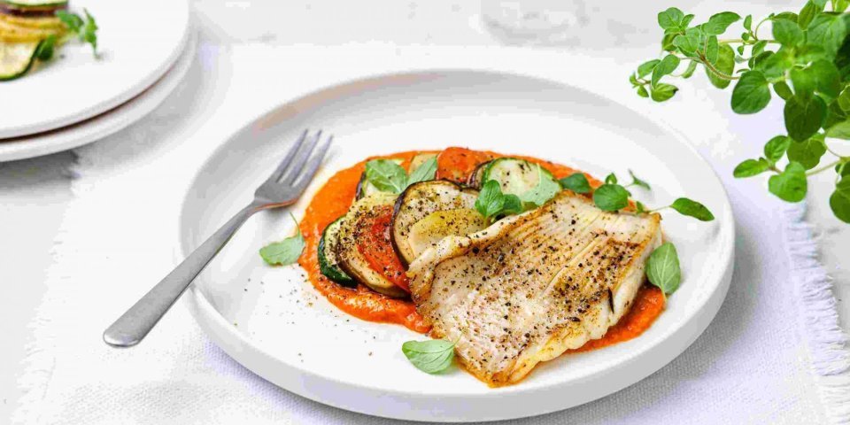 Gebakken rog met ratatouille en paprikasaus Gebakken rog met ratatouille en paprikasaus geserveerd op een wit groot bord. Het vork ligt op het bord en verse kruiden staan langs deze hoofdmaaltijd.