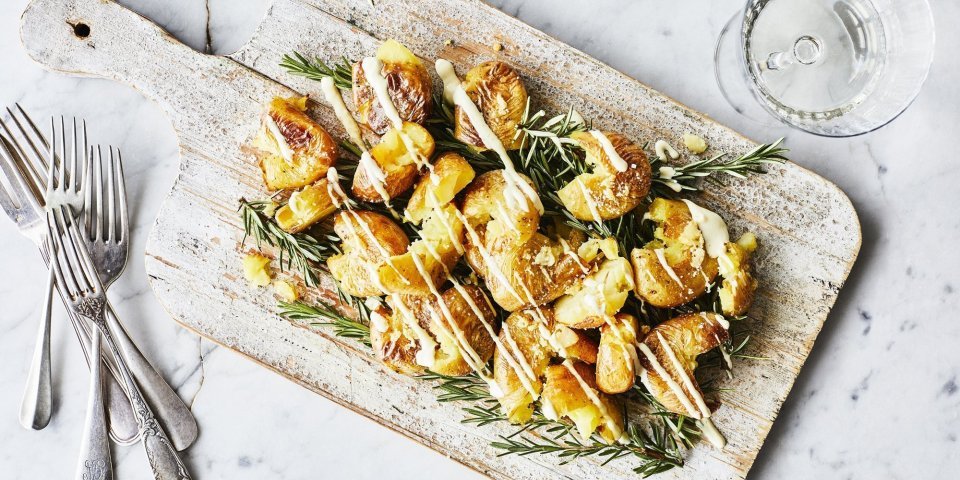 Gecrushte aardappelen met picklessausje Eens een patatje in een feestjas op tafel? Serveer deze ovenaardappeltjes en ontvang veel "mmmmm-lekker complimentjes". Deze gecrushte aardappeltjes zijn geserveerd op een houten plank met een heerlijk picklessausje.