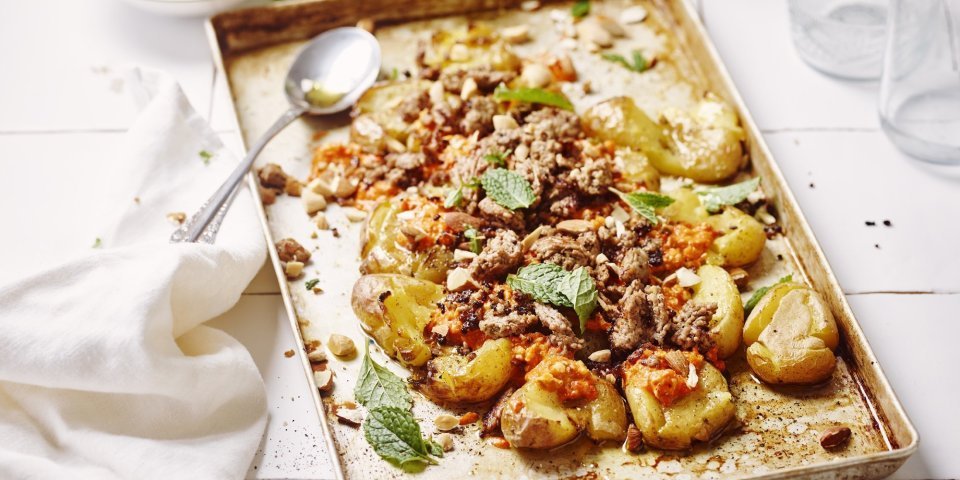 Op een ovenschaal liggen krokante crushed potatoes met homemade romesco saus bovenop en afgewerkt met lamsgehakt. De overige romesco saus staat bovenaan voor wie wat extra wil. Er ligt een witte servette op het aanrecht van witte Marokaanse tegeltjes. 