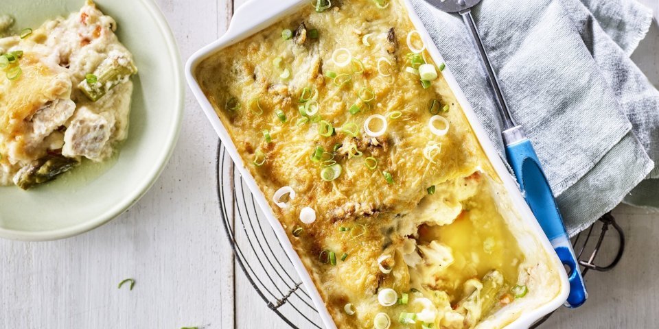Gegratineerd vispannetje met Brugge kaas, asperges en venkel Het vispannetje staat op een metalen onderlegger op een witte houte tafel. De schepper ligt naast de schotel waaruit al 1 portie is uitgeschept in een lichtgroen bordje.
