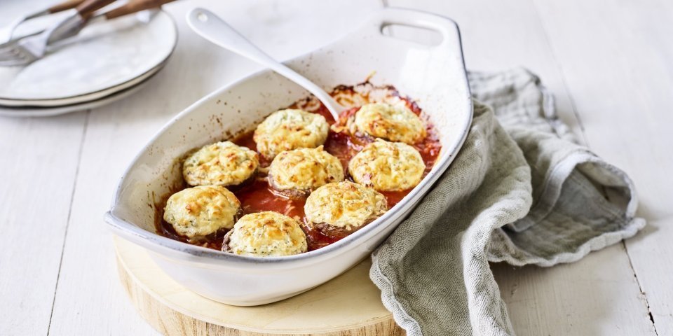 Witte ovenschotel staat op een houten snijplankje op tafel. De ovenschotel is gevuld met de rode tomatensaus waarin de champignons mooi in geschikt zijn. Achteraan de foto zie je bordjes staan met bestek om aan de slag te gaan. 