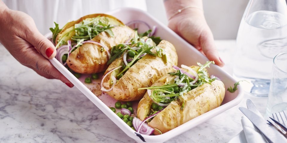 Gepofte aardappel met pesto, erwtjes en rucola 3 gepofte aardappelen met pesto, erwtjes en rucola liggen in een witte ovenschaal. De vrouw wil de ovenschaal plaatsen op tafel.