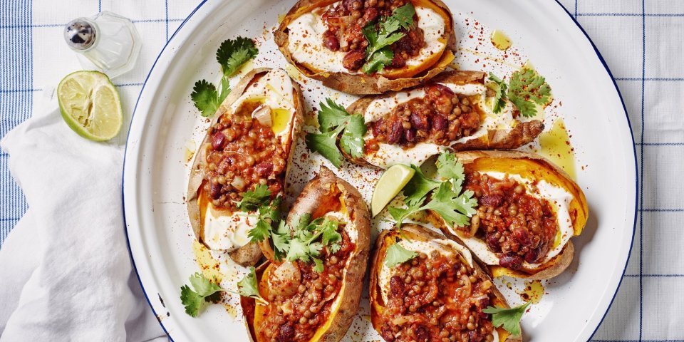 Een ronde ovenschaal met de gepofte zoete aardappelen staat op tafel. De gepofte aardappelen zijn gevuld met yoghurt en hierbovenop de chili sin carne, ze zijn afgewerkt met enkele blaadjes koriander en partjes limoen. De witte ovenschaal heeft een blauw randje en staat op een blauw geruite handdoek.