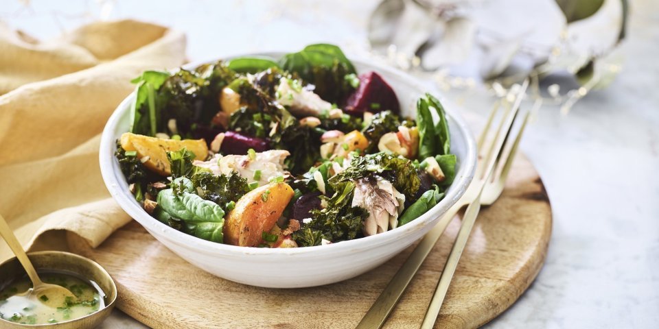 Een salade van bietjes, boerenkool en spinazie afgewerkt met noten en makreel. Deze feestsalade staat op een houten plank met de dressing in een apart gouden potje.