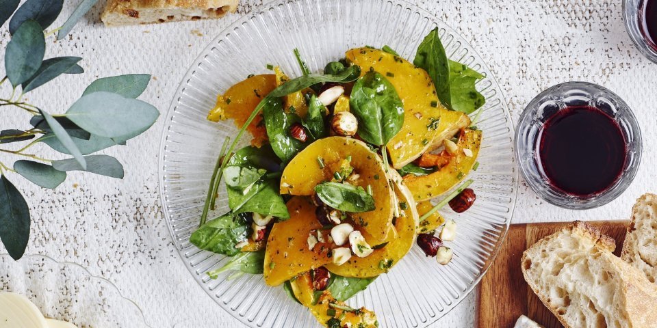 Een glazen bordje staat op tafel met geroosterde partjes pompoen. De salade krijgt een mooi groen kleurtje door de verse spinazie, hij is afgewerkt met enkele hazelnoten. Op en top een herfstsalade boordevol smaak.