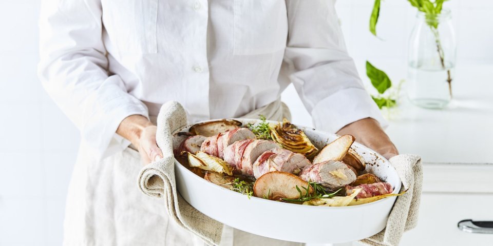 Gevuld varkenshaasje met Belgische smeltkaas, peer en venkel Een vrouw met witte linnen hemd en beige keukenshort draagt een witte ovenshotel waarin het varkenshaasje in ligt. De keuken achter zich is wit van kleur. Er staat een kastje naast haar waarop een vaasje staat met witte bloem in.