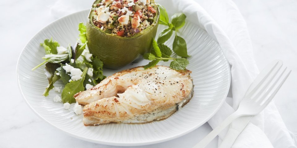 Een gebakken moot van schelvis ligt op een wit bord. Hij is geserveerd met een gevuld groene paprika. Deze is opgevuld met quinoa, tomaten- en komkommerstukjes en geitenkaasblokjes.