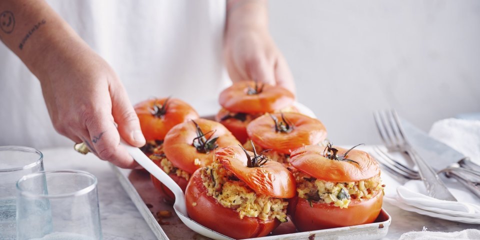 Zes gevulde rode tomaten in een ovenschaal zijn aan tafel gebracht door een vrouw in witte t-shirt. Ze schept alvast eentje op voor zichzelf.