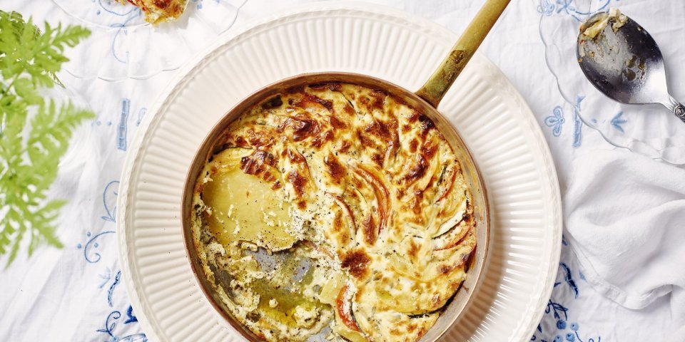 Een heerlijke gratin met groenten en kaas klaargemaakt in een ovenbestendige pan staat in het midden van de tafel. Er is al een portie uitgeschept op een bordje. Op tafel ligt een wit tafelkleed met enkele geborduurde blauwe bloemen. 