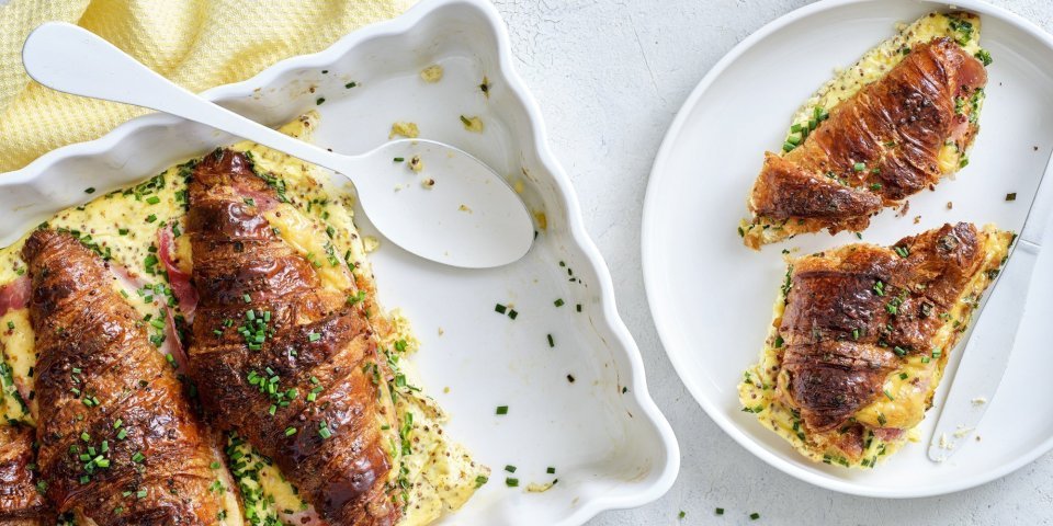 Hartige croissants uit de oven met ham, kaas en mosterd Er staat een witte ovenschotel op tafel gevuld met croissants. Ze zijn doormidden gesneden, bestrijkt met mosterd en belegd met ham en kaas. Daarna is een beslag toegevoegd en werden ze 10 minuten in de oven geplaatst. Om vingers en duimen van af te likken, al zeg ik het zelf.